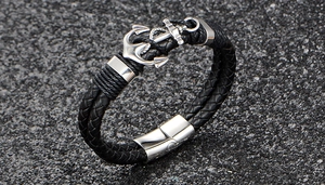 Detail of black braided leather bracelet featuring a silver anchor charm on stone surface