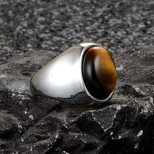 Mens stainless steel ring featuring a large oval tiger eye stone on a dark rock background