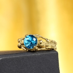 Anello gotico dorato con teschio e gemma blu centrale su scatola nera testurizzata