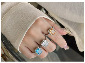 Three halo radiant cut gemstone rings in champagne, white and blue on a model's hand