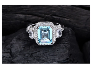 Macro shot of emerald-cut blue crystal ring with diamond halo on dark coal background