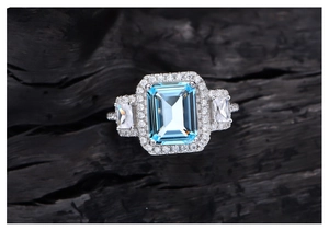 Macro detail of silver ring with emerald cut light blue stone and diamond halo on dark wood