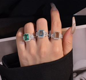 Three emerald-cut colored rings with halo diamonds worn against a magazine background