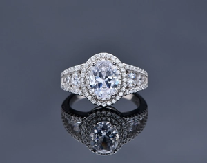 Macro shot of oval-cut diamond ring with double halo and pave band on dark mirror