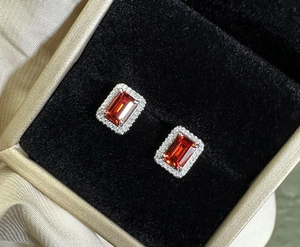 Macro detail of emerald-cut orange-red ruby earrings with white diamond halo on velvet