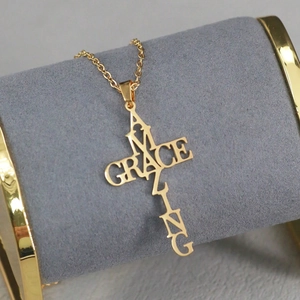 Collier croix Amazing Grace en acier inoxydable doré exposé sur un support en velours gris