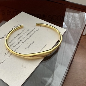 Bracciale rigido dorato opaco dal design organico e aerodinamico su un biglietto poetico