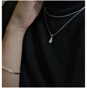 Detail shot of a minimalist silver teardrop pendant and layered chains on a black sweater
