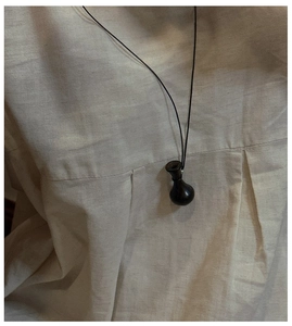 Dettaglio del ciondolo a vaso in legno scuro su cordino nero indossato su camicia di lino beige