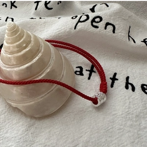 Détail du bracelet tressé rouge avec perle en argent martelé posé sur un coquillage blanc
