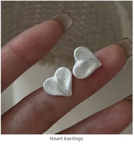 Macro shot of silver heart stud earrings with a brushed matte finish held by fingers