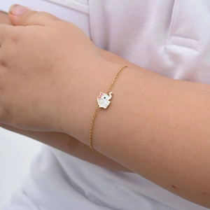 Detalle de una pulsera dorada infantil con dije de elefante de esmalte blanco y lazo rosa