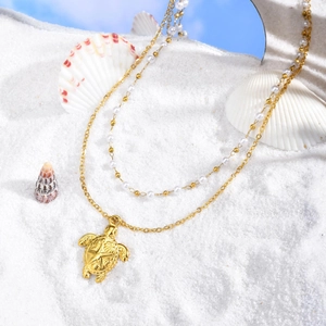 Collier double rang doré avec pendentif tortue et perles sur un fond de plage ensoleillé