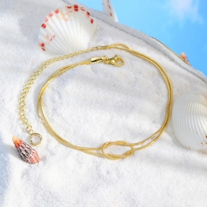Gold double-layered snake chain anklet with a knot design on white beach sand