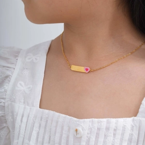 Détail d'un collier doré avec plaque courbée et cœur rose porté par un mannequin