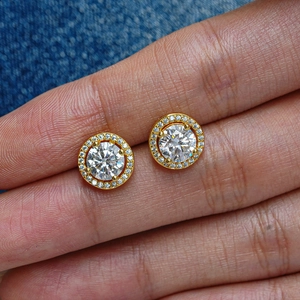 Classic round moissanite halo stud earrings with gold finish held by hand on denim