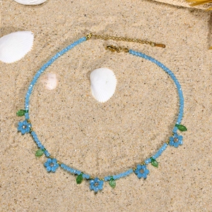Collier en perles bleues avec accents floraux et feuilles vertes sur fond de sable ensoleillé