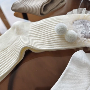 Detail view of cream mid-calf ribbed socks featuring white lace trim and plush pom-poms