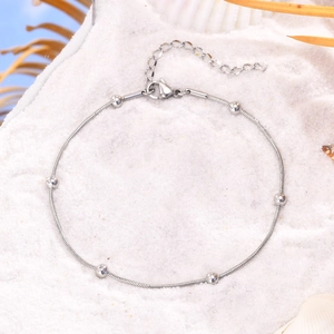 Detail of a golden stainless steel beaded anklet on white sand with sea shells