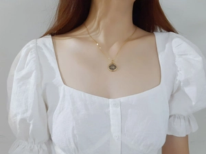 Détail du collier pendentif œil porte-bonheur doré porté sur un chemisier à manches bouffantes
