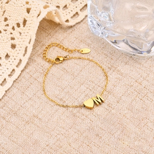 Vue détaillée du bracelet en acier doré avec lettre M et pendentif cœur sur tissu beige