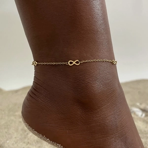 Close-up of a gold infinity charm anklet worn on skin against a beach background