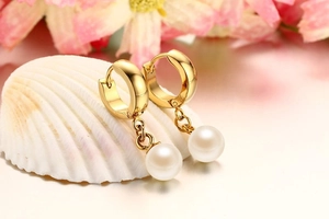 Detailed close-up of gold huggie earrings with round white pearl pendants on a shell