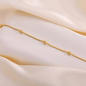 Detail of golden stainless steel snake chain anklet with beads on white silk fabric