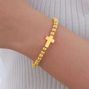 Gros plan d'un bracelet en perles dorées polies avec un pendentif croix minimaliste au poignet