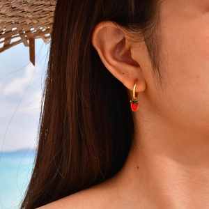 Gros plan d'une femme portant des créoles fraise en émail doré sur une plage d'été ensoleillée
