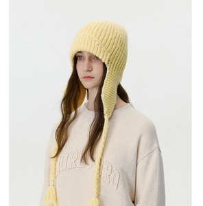 Close-up of cable knit earflap beanie in pale yellow with braided tassels and pom-poms