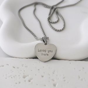 Détail macro du pendentif coeur gravé 'Love you more' avec sa fine chaîne box
