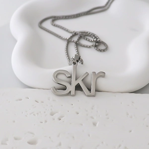Detalle del colgante de letra SKR plateado en cadena fina sobre fondo artístico blanco