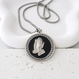Macro detail of silver necklace pendant featuring a 3D praying hands motif and Greek key border