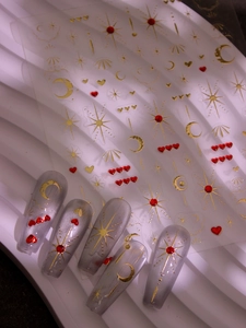 Detail of star and moon gold nail stickers applied on cat-eye grey nails with red hearts