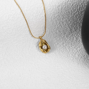 Elegant gold-tone shell-shaped pearl pendant necklace with thin snake chain on textured white background