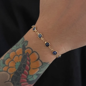 Dettaglio di un bracciale dorato con occhio di Allah e ciondoli blu su un polso tatuato