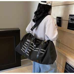 Rear view of a black canvas travel tote bag with white lettering on wooden shelf background