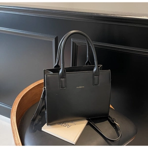 Close-up of black matte square handbag with gold logo detail on a leather chair