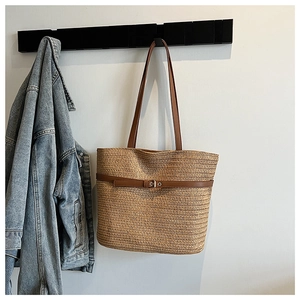 Detail of woven straw tote bag featuring brown leather straps and buckle on white wall
