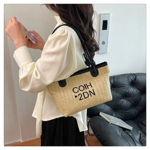 Woven straw tote bag with black leather straps and embroidered letters worn by a model