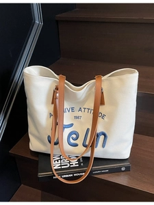 Off-white canvas tote bag with blue embroidery and brown leather handles on wooden steps