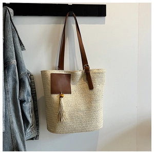 Detail of a beige woven straw tote bag with brown leather straps and wooden bead tassel