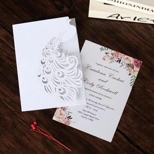 Intricate laser-cut white peacock wedding invitation with floral insert on wood background
