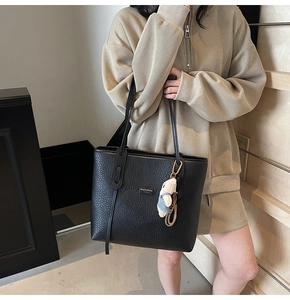 Detail of black pebbled leather tote bag featuring metal logo and white plush bear charm
