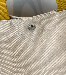 Détail du sac cabas en toile montrant la texture beige, le bouton pression et la sangle