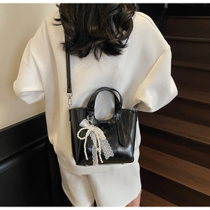 Detail of black glossy handbag with lace pearl bow charm worn by a model in white hoodie