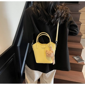 Bright yellow bucket bag with brown teddy bear charm shown in a lifestyle street setting