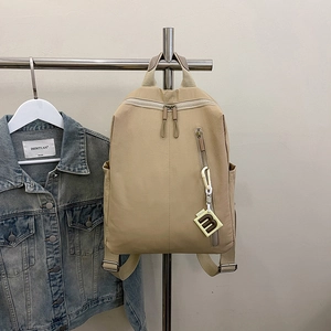 Detail view of beige nylon backpack featuring dual zippers and a lettered square pendant