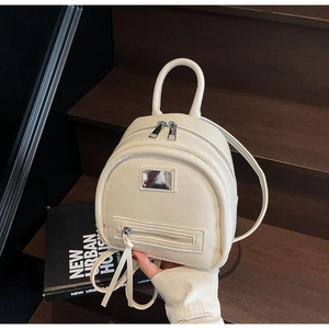 Detail view of cream mini backpack with dual zippers and metal plaque on wooden stairs